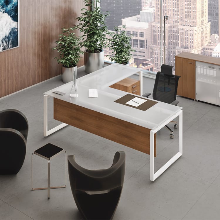 A modern office showcases a Cubiko Managerial white and wood L-shaped desk paired with a sleek black chair. Two stylish black lounge chairs and a small side table create an inviting seating area near expansive windows that offer sweeping city views, while two potted plants by the window add a touch of greenery. Studio Office Cyprus