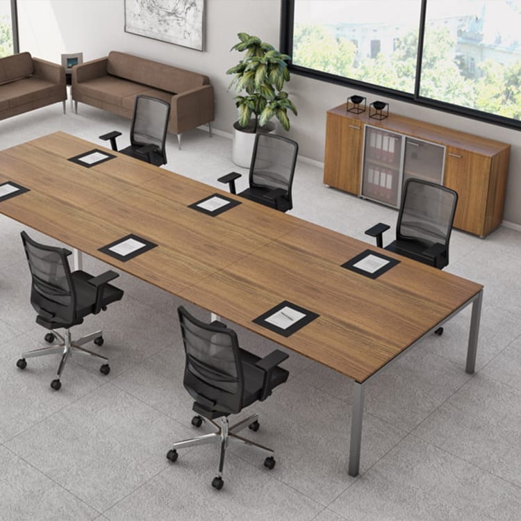 A modern office space showcases the I-Work Conference table surrounded by six black mesh chairs. Brown couches and sleek cabinets add warmth, while a large plant brings a touch of greenery. Sunlight streams through a window with city views, and documents are neatly arranged on the conference table. Studio Office Cyprus