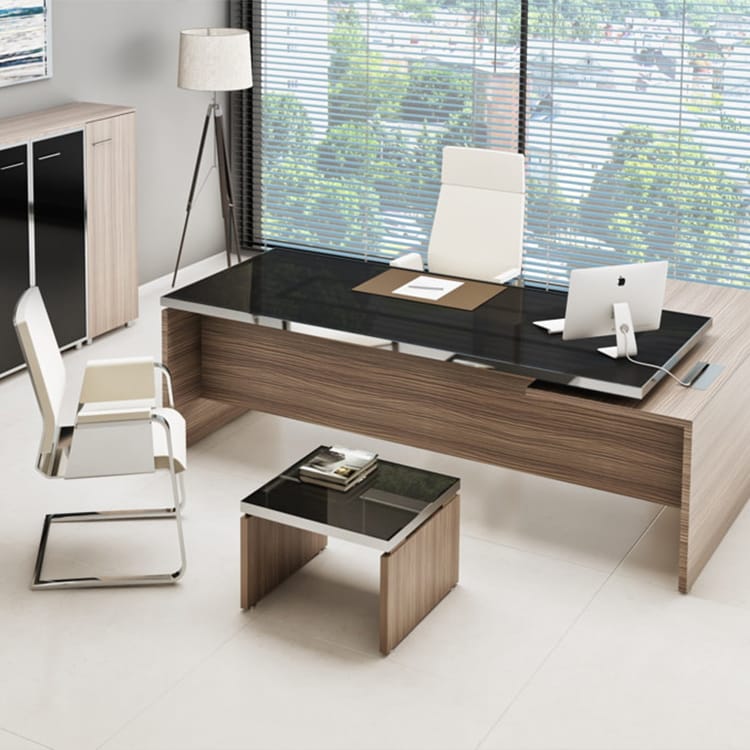 A contemporary office showcases the Manager EVO Executive wooden desk with a sleek glass top, paired with white leather chairs. A computer and stylish lamp sit atop the desk, while expansive windows with blinds offer views of vibrant greenery outside. Minimalist decor creates an elegant, modern atmosphere—perfect for the discerning manager who values sophisticated style. Studio Office Cyprus