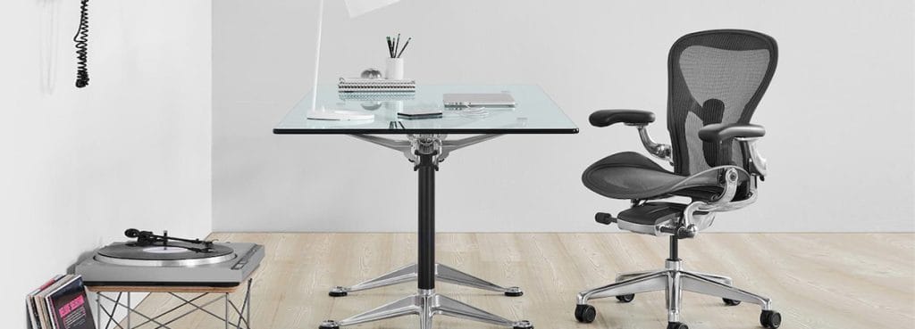 Ergonomic graphite Aeron Chair with a mesh back and adjustable arms near a glass top desk.